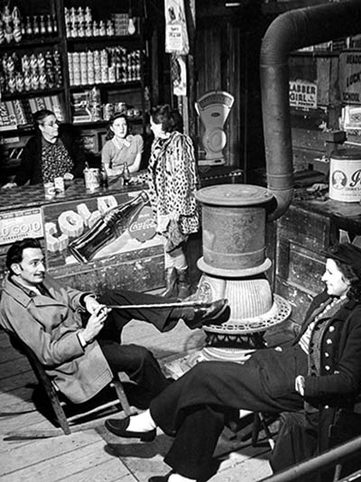 Surrealist artist Salvador Dali (L) w. wife (R), warming themselves around a pot-bellied stove as their host Mrs. Phelps Crosby stands at counter emblazoned w. Coca Cola ad, as she shops at Safeway Grocery store. (Photo by Eric Schaal/Pix Inc./The LIFE Images Collection/Getty Images)